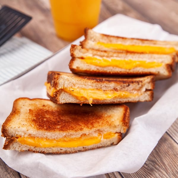 Three grilled cheese sandwiches with melted cheddar, served on white paper with an orange drink.
