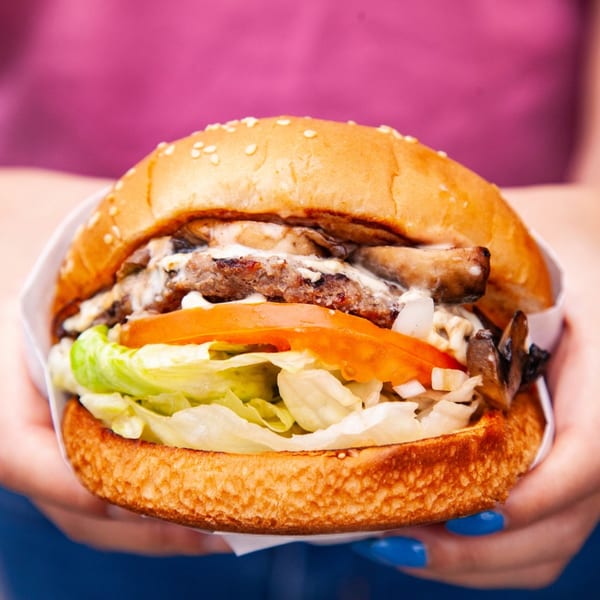 A person holds a John's Place burger with lettuce, tomato, onions, and mushrooms.