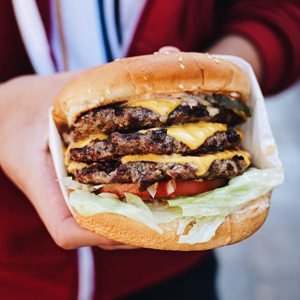 A hand holds a John's Place triple cheeseburger with lettuce, tomato, and onions.