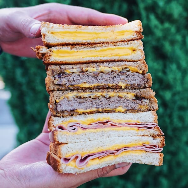 Hands holding a towering sandwich with layers of grilled cheese, burger patties, and ham.