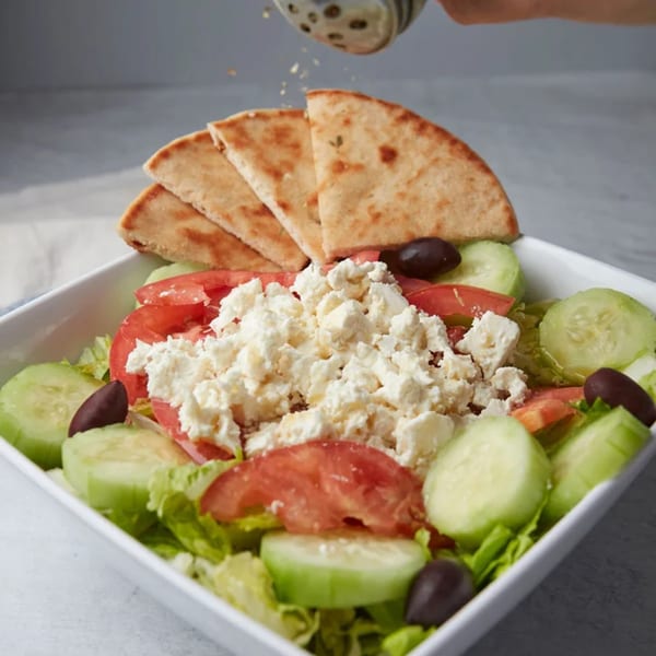 John's Place Greek salad with feta, tomatoes, cucumbers, olives, and pita bread, being seasoned.