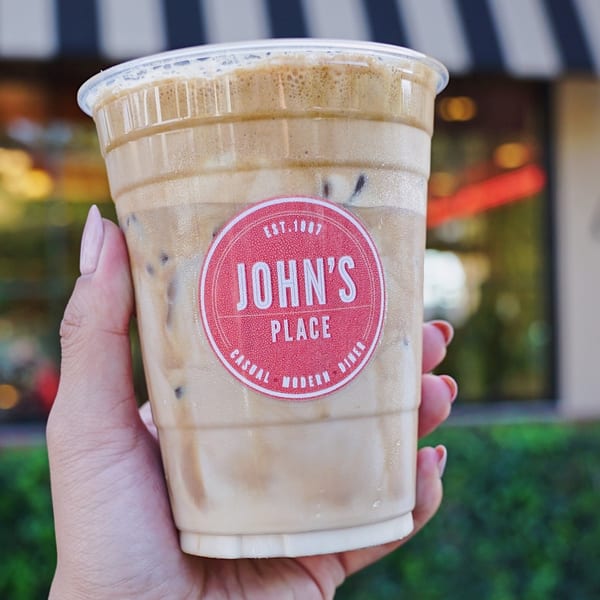 Hand holding a John's Place iced coffee with a red logo sticker, outdoors.