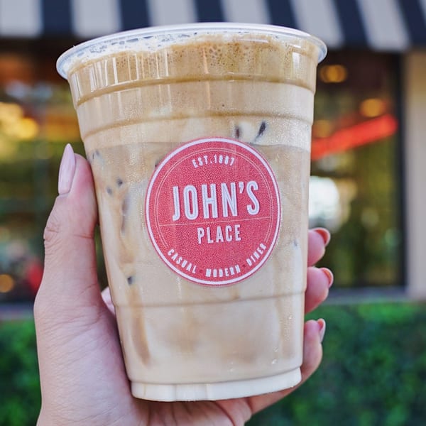 Hand holding an iced coffee from John's Place with a red logo sticker on the cup.