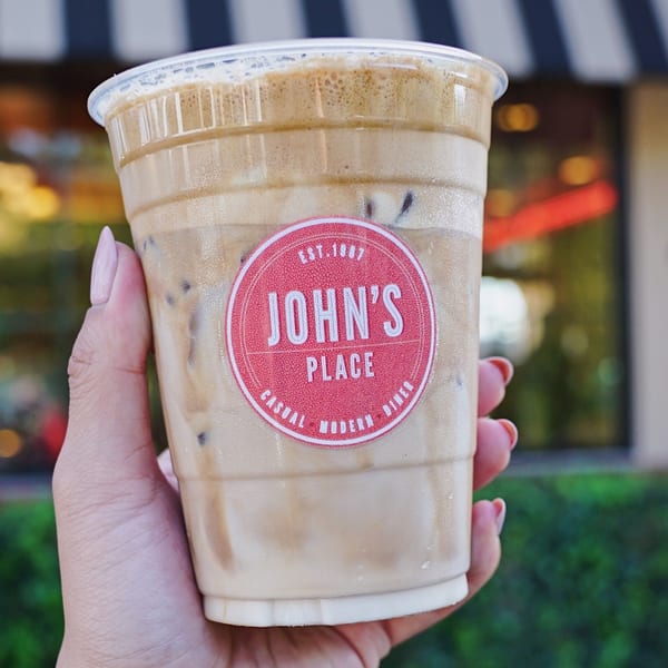 Hand holding an iced coffee with a red John's Place logo sticker on the cup.