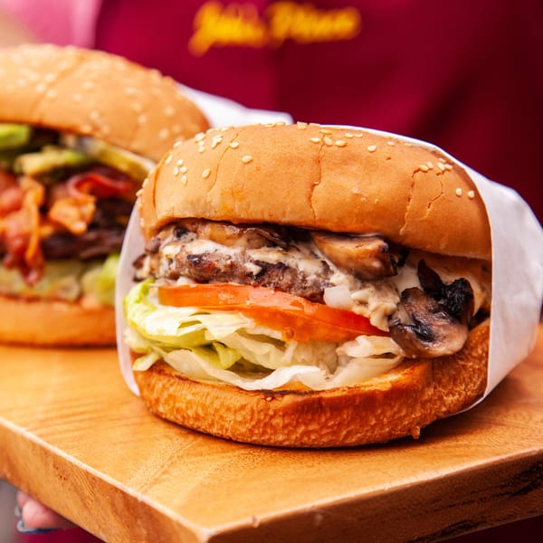 Two John's Place burgers on a wooden board, one with mushrooms, the other with bacon and avocado.