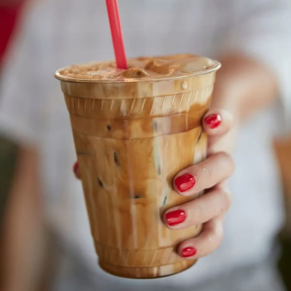 A hand with red nail polish holds a plastic cup of iced coffee with a red straw.