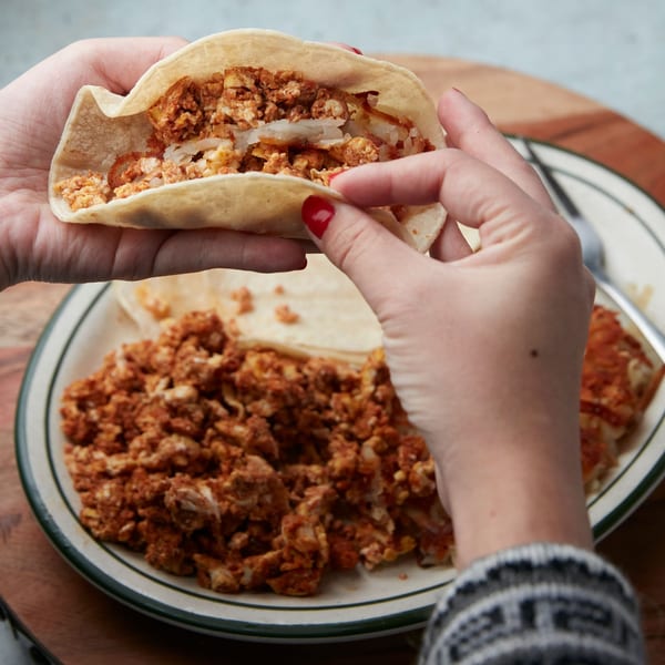 Hands holding a taco filled with scrambled eggs and potatoes, with more on a plate.