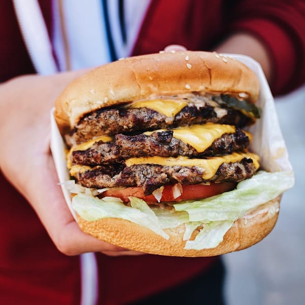 A hand holding a John's Place triple cheeseburger with lettuce, tomato, and onions.
