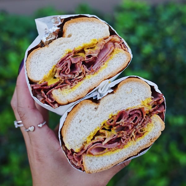 A hand holds a John's Place pastrami sandwich, cut in half, with mustard on white bread.
